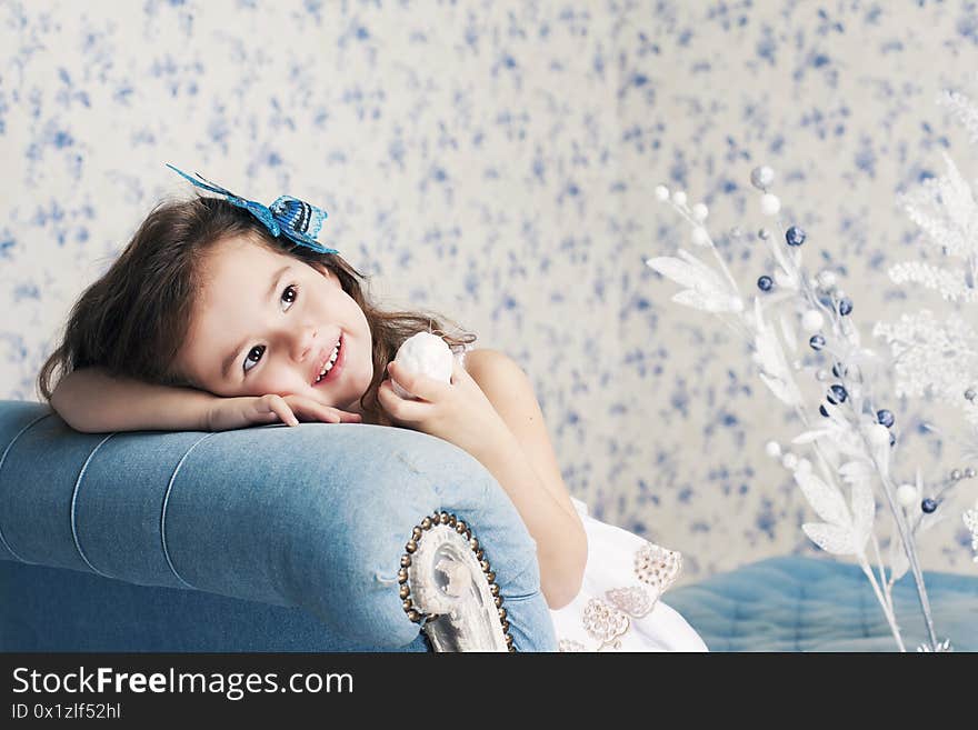 Cute smiling girl in white dress sitting on blue sofa with Christmas decorations. Holidays, New Year celebration concept.