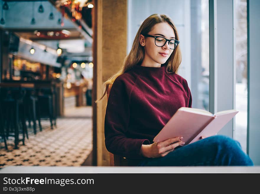 Successful female student wear in spectacles for vision correction enjoying literature at university cafeteria, caucasian intelligent hipster girl casual dressed reading positive book indoors. Successful female student wear in spectacles for vision correction enjoying literature at university cafeteria, caucasian intelligent hipster girl casual dressed reading positive book indoors