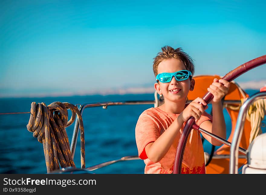 A handsome boy of 9-10 years old at the helm of a yacht in bright orange clothes, blue glasses against a blue sky. Sea vacation concept, sailing regatta. Traveling with children