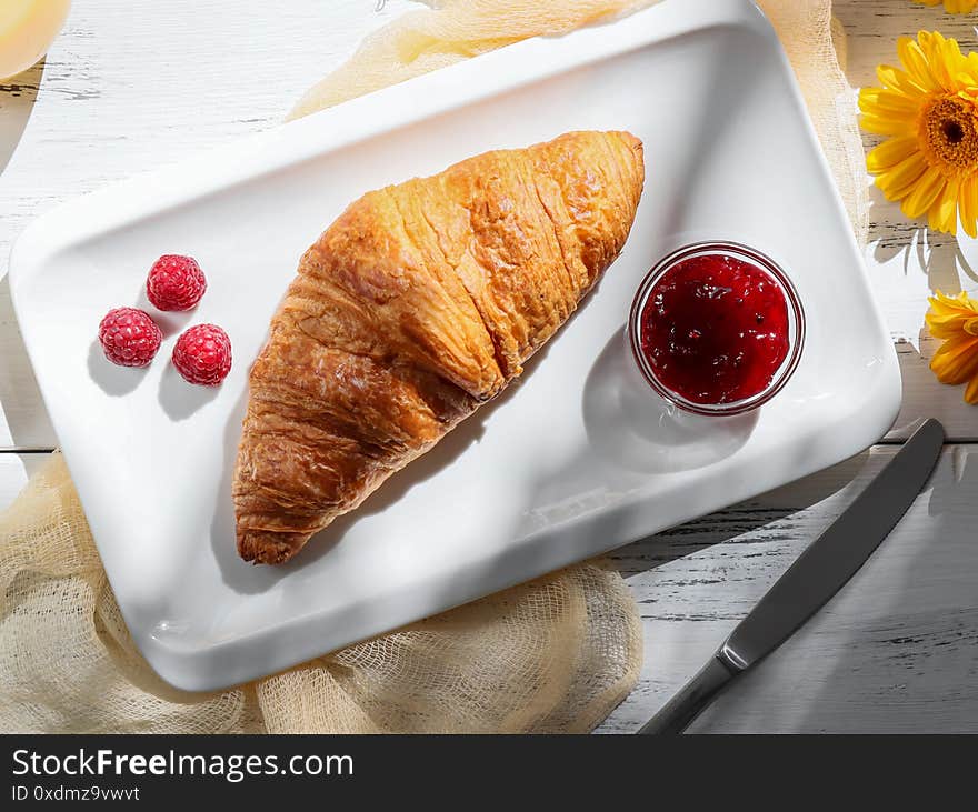 One croissant with berry jam on a white rectangular plate lies on a white wooden table with an orange gerbera and fresh orange juice. One croissant with berry jam on a white rectangular plate lies on a white wooden table with an orange gerbera and fresh orange juice.