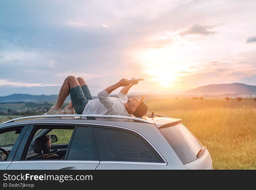 Mature age Man lying on car roof and reading the paper bestseller book. He stopped his auto on high grass meadow with beautiful