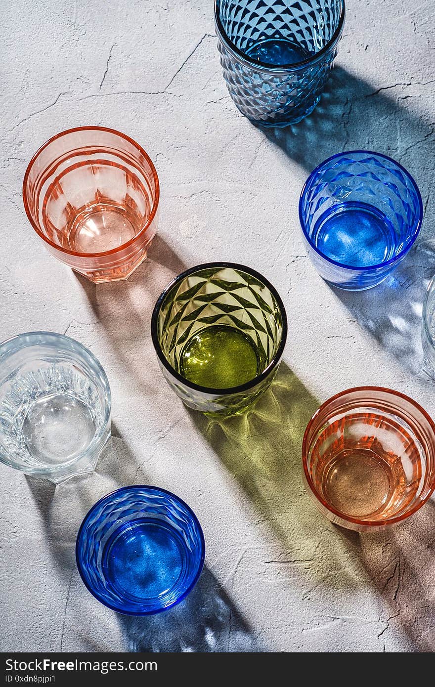 Colorful faceted and geometric drink glass, group of empty green, red, blue and transparent drinkware for beverage on stone concrete background, angle view