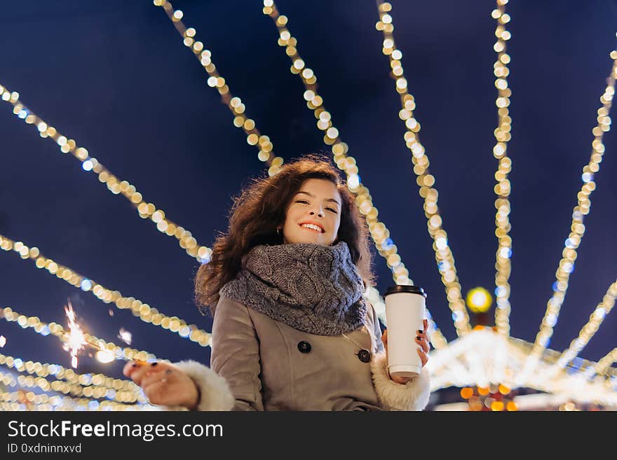 Girl Having Fun With Sparklers On Christmas Decoration Lights Street. Young Happy Smilin Woman Wearing Stylish Knitted Scarf, Coat