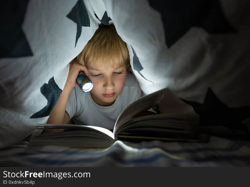 A little boy reads a book with a flashlight under the covers at night.