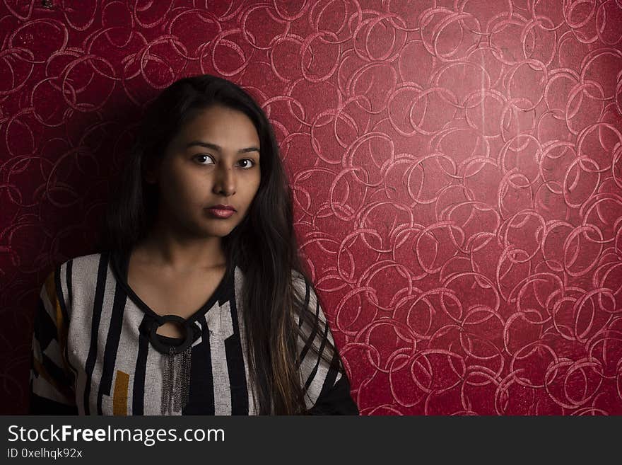 Fashion portrait of an Indian Bengali beautiful and young girl in western dress standing in front of a red textured wall.