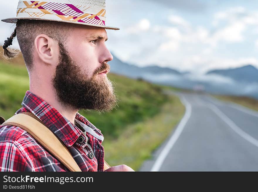 Handsome young man in a straw hat and a plaid shirt, stands on the road, against the backdrop of beautiful mountains at sunset. The concept of freedom and hitchhiking.