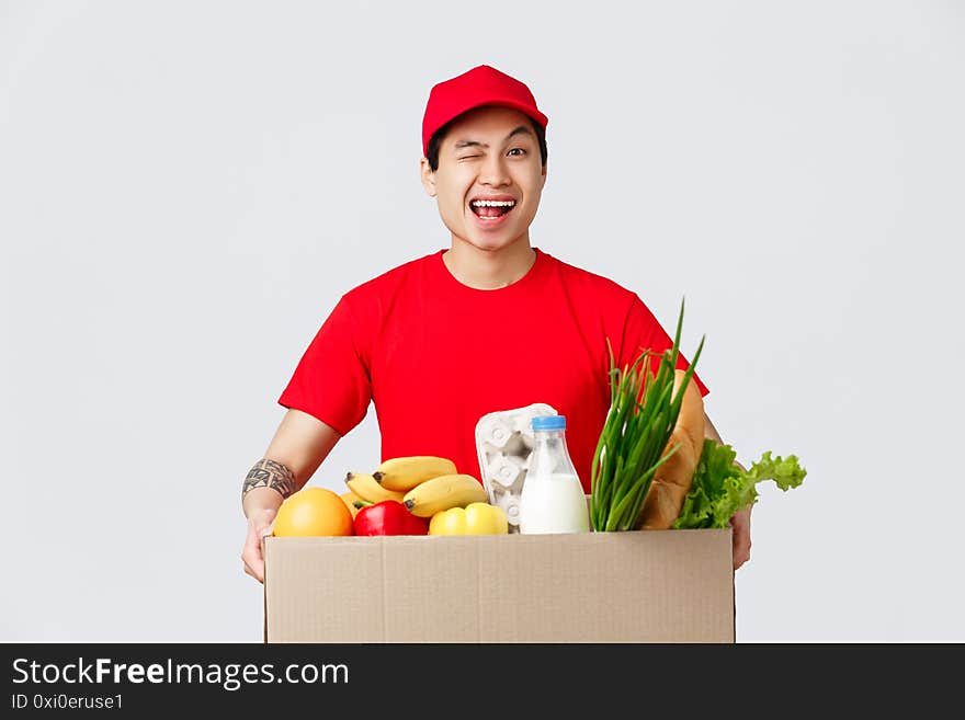 Online shopping, food delivery and internet stores concept. Friendly smiling asian courier in red cap and t-shirt, holding package with grocery deliver, wink customer, give client product packages.