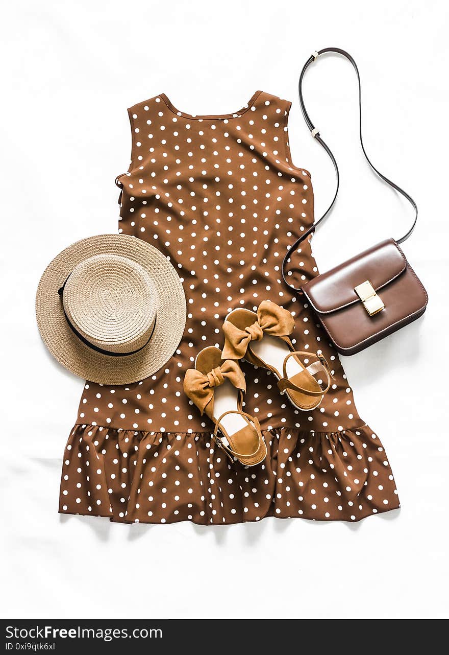 Summer women outfit look - brown polka dot sleeveless dress, suede sandals, leather crossbody bag, sunglasses and hat on a light background, top view.