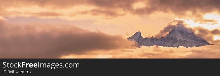 Foggy clouds and sunshine in evening overcast sky view. Famous Germany Alps Watzmann Mountain top silhouette in clouds. Natural weather and climate sky background scene. Foggy clouds and sunshine in evening overcast sky view. Famous Germany Alps Watzmann Mountain top silhouette in clouds. Natural weather and climate sky background scene