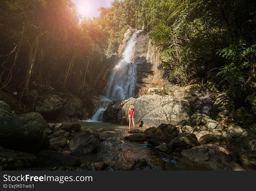 Beautiful young slim blonde woman with curly hair, wearing in red swimsuit is enjoy in lagoon of huge tropical waterfall