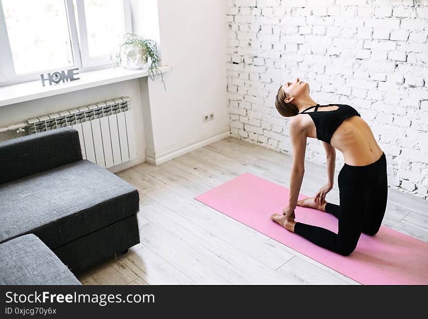 Bendy girl stands on her knees and touching her heels in back bent pose. A young girl is doing her home workout near a bright window on a fitness mat. Bendy girl stands on her knees and touching her heels in back bent pose. A young girl is doing her home workout near a bright window on a fitness mat