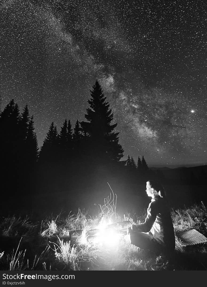 Side view of female tourist having a rest by campfire on background of spruce forest under starry sky on which milky way is visible. Bonfire with a big flame under the night sky. Monochrome image. Side view of female tourist having a rest by campfire on background of spruce forest under starry sky on which milky way is visible. Bonfire with a big flame under the night sky. Monochrome image