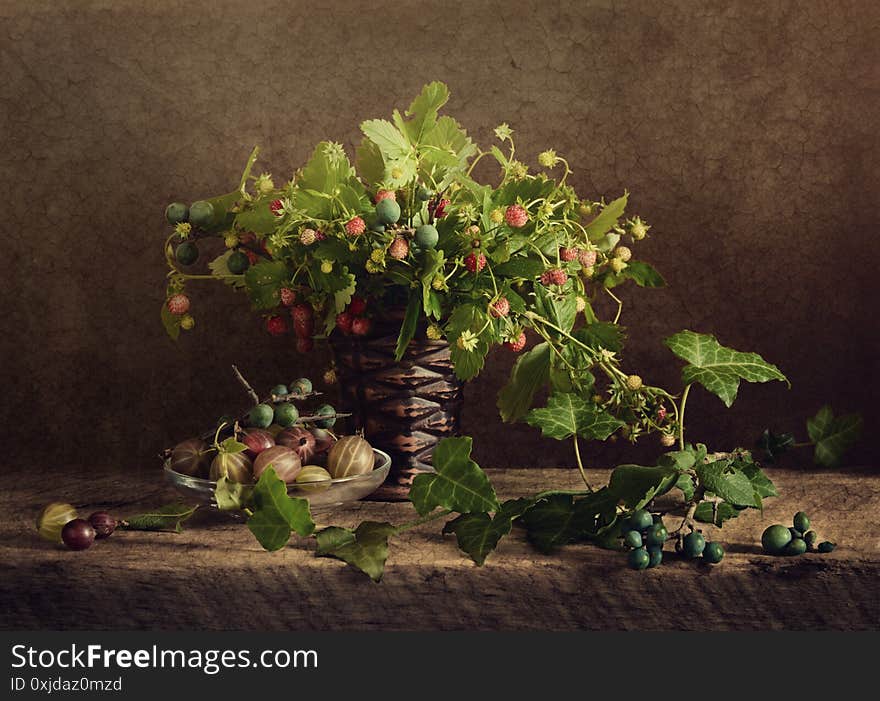 Wild plants still life. Red strawberry berry bouquet in vase and green leaves on dark brown vintage background