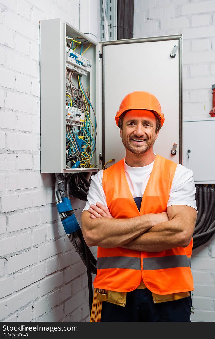 Handsome electrician in hardhat and safety vest smiling at camera near electrical distribution box