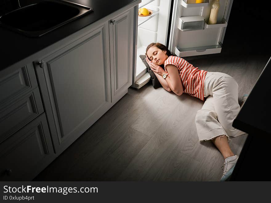 High angle view of attractive girl sleeping on pillow near open refrigerator in kitchen at night