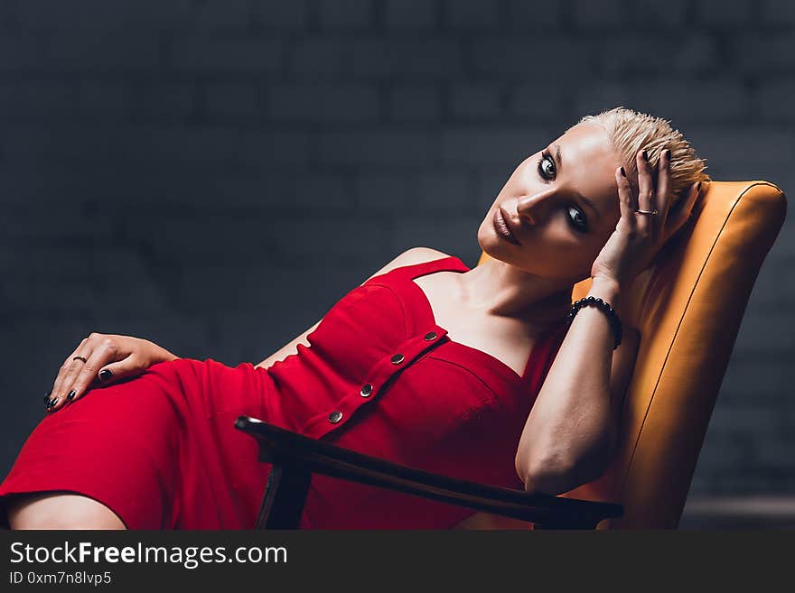 Young woman with blonde hair and a short haircut in a red evening dress lies on the chair.