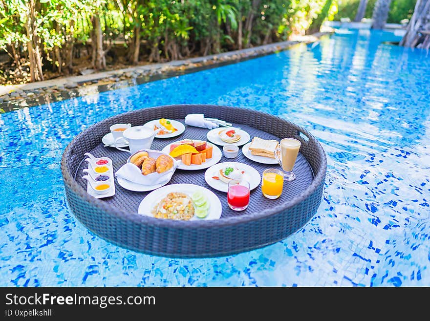 Floating breakfast set in tray with fried egg omelette sausage ham bread fruit milk juice coffee and other around swimming pool