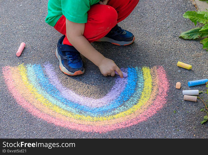 A boy draws a rainbow on the asphalt with crayons . A child draws on the asphalt . Children`s drawing. Walking in the city with a child. Summer in the city. Crayons for drawing. Article about childhood