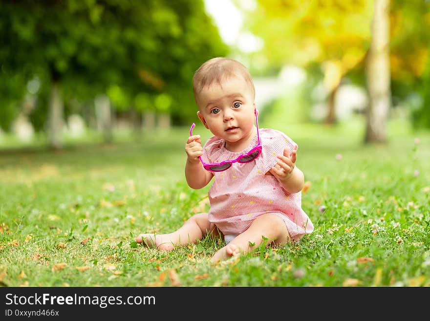 Little baby girl 7 months old sitting on the green grass in a pink bodysuit and bright glasses, walking in the fresh air.