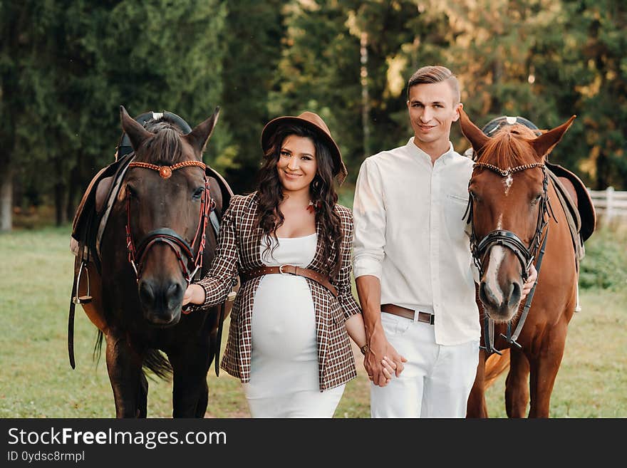 A Pregnant Girl In A Hat And Her Husband In White Clothes Stand Next To Horses In The Forest In Nature.Stylish Pregnant Woman With