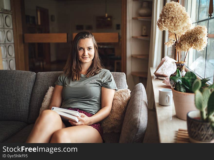 Portrait of attractive young woman holding book while relaxing on couch in living room at home. High quality photo