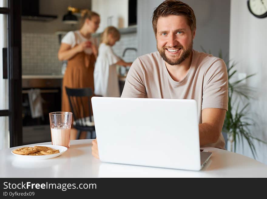 Happy positive guy at freelance during quarantine, young male sit using laptop, woman and child girl in the background. Happy positive guy at freelance during quarantine, young male sit using laptop, woman and child girl in the background