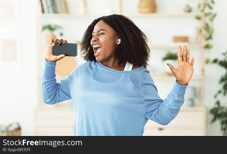 Carefree Singer. Portrait of african lady singing and using cell phone as mic, dancing and wearing earbuds. Carefree Singer. Portrait of african lady singing and using cell phone as mic, dancing and wearing earbuds