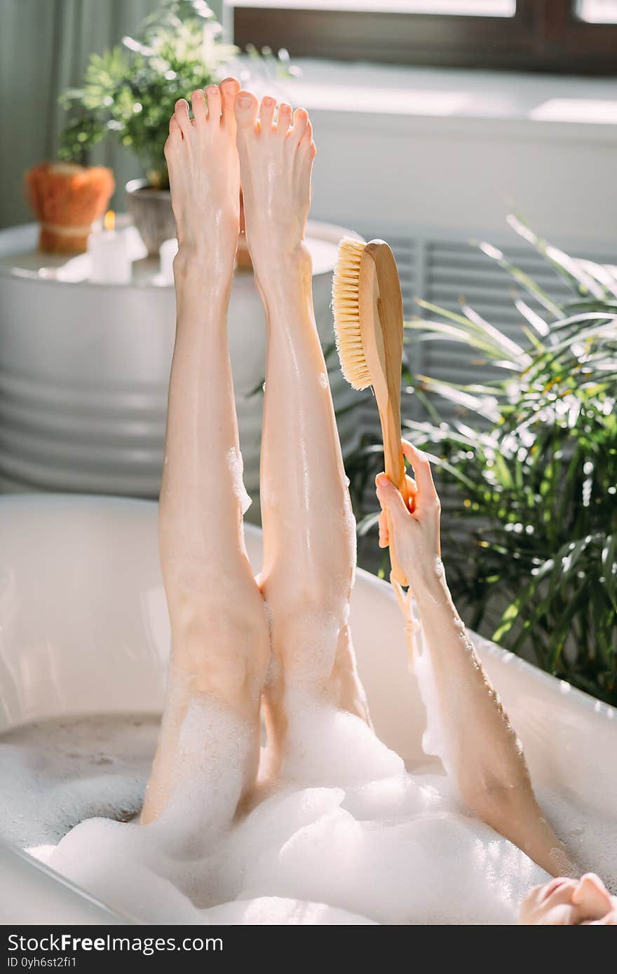 Cropped shot of female slim legs raised in air. Bathing unrecognizable woman massaging her slender long perfect legs with brush after depilation in bathroom. Cropped shot of female slim legs raised in air. Bathing unrecognizable woman massaging her slender long perfect legs with brush after depilation in bathroom