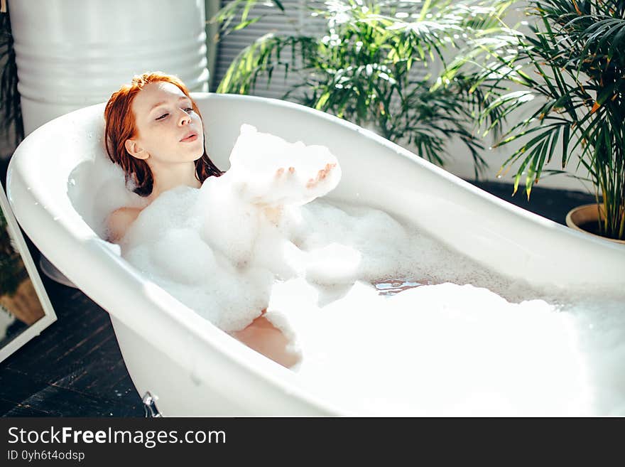 Playful young caucasian woman enjoying foam bath at home. Resting, relaxing, blowing on a cloud of foam. Playful young caucasian woman enjoying foam bath at home. Resting, relaxing, blowing on a cloud of foam