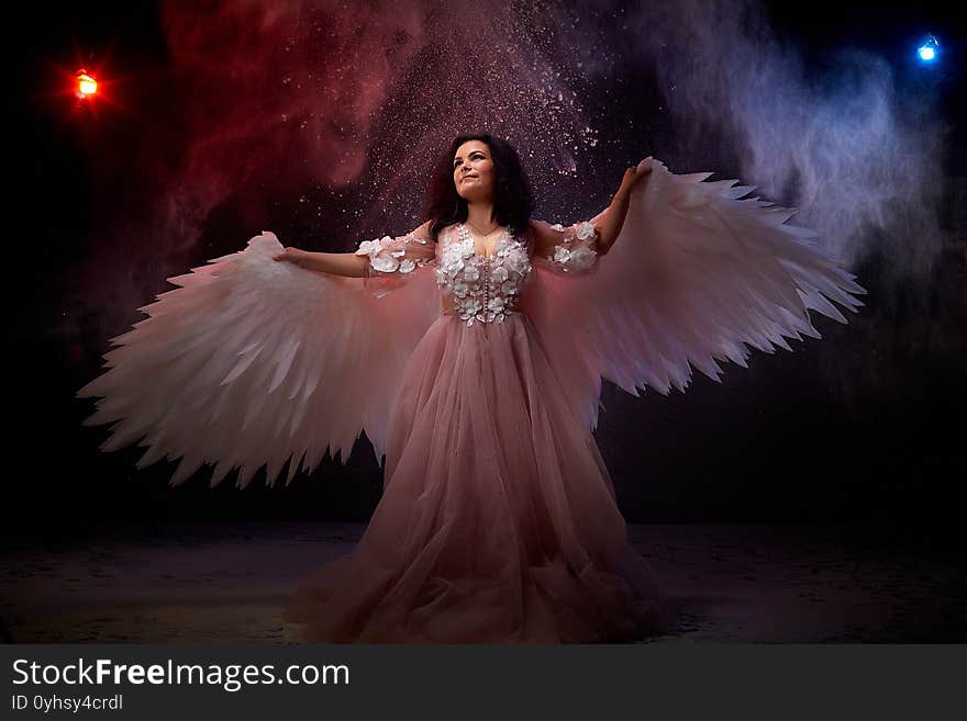 Girl with white wings posing in dark black studo during photoshoot with flour or dust and light. White angel in dark space. Concept struggle between good and evil