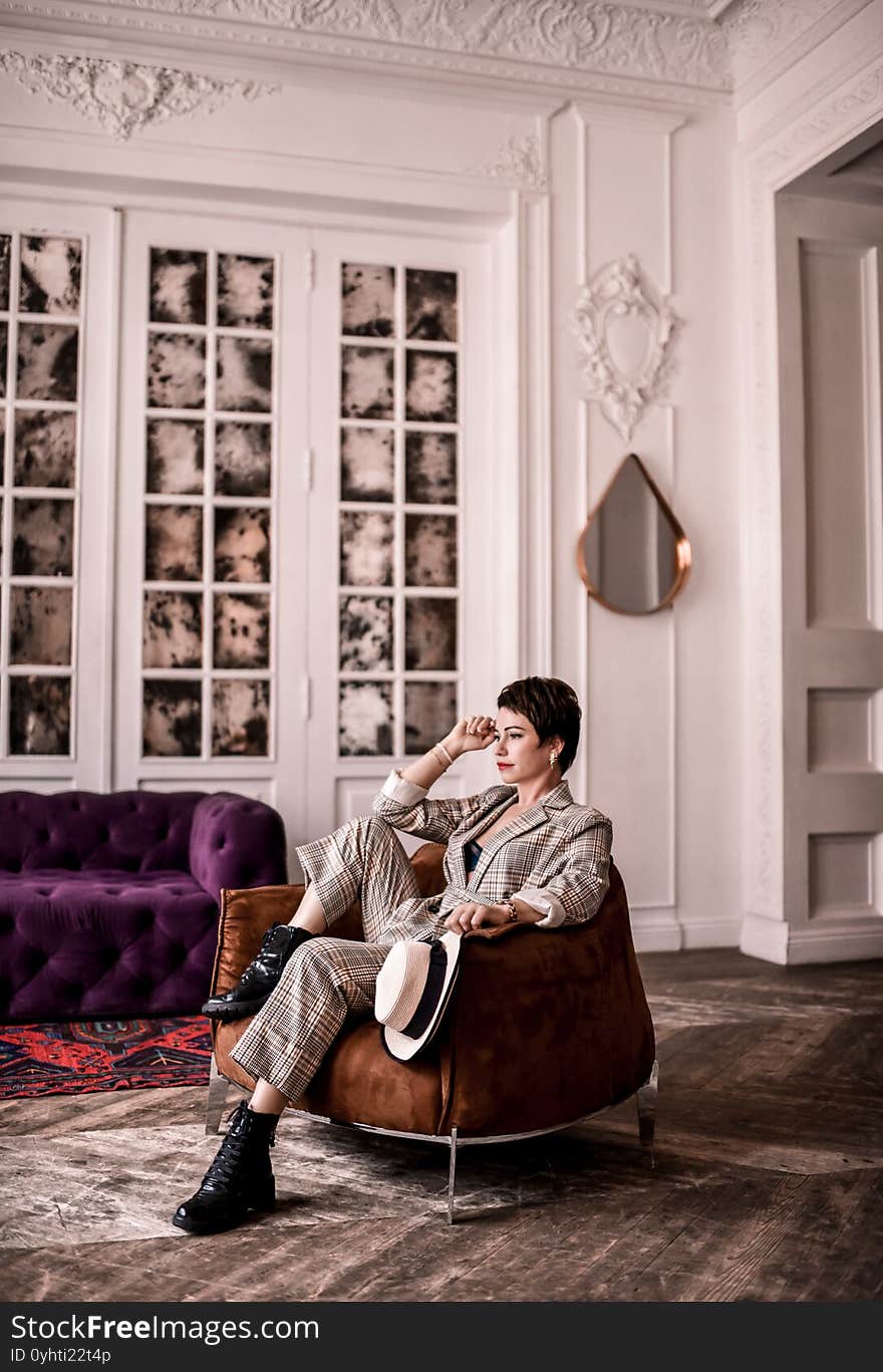 Calm short haired brunette woman in stylish business plaid pantsuit with rolled up sleeves sits in armchair holding hand at head and think. Business elegance concept. Calm short haired brunette woman in stylish business plaid pantsuit with rolled up sleeves sits in armchair holding hand at head and think. Business elegance concept