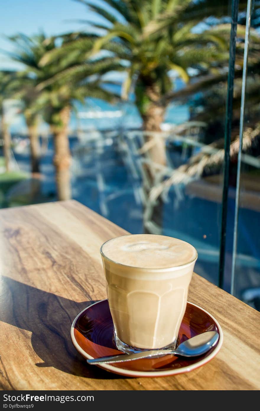 A glass of coffee latte on a wooden table with tropical nature background - palms and the ocean on a sunny day