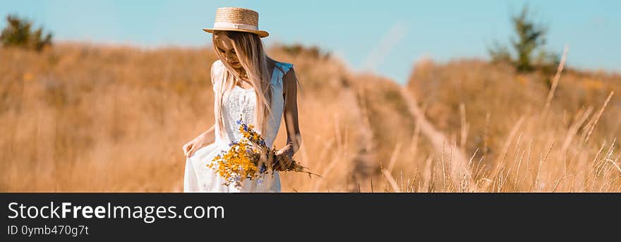 Horizontal image of blonde woman in straw hat touching white dress while standing in field with bouquet of wildflowers