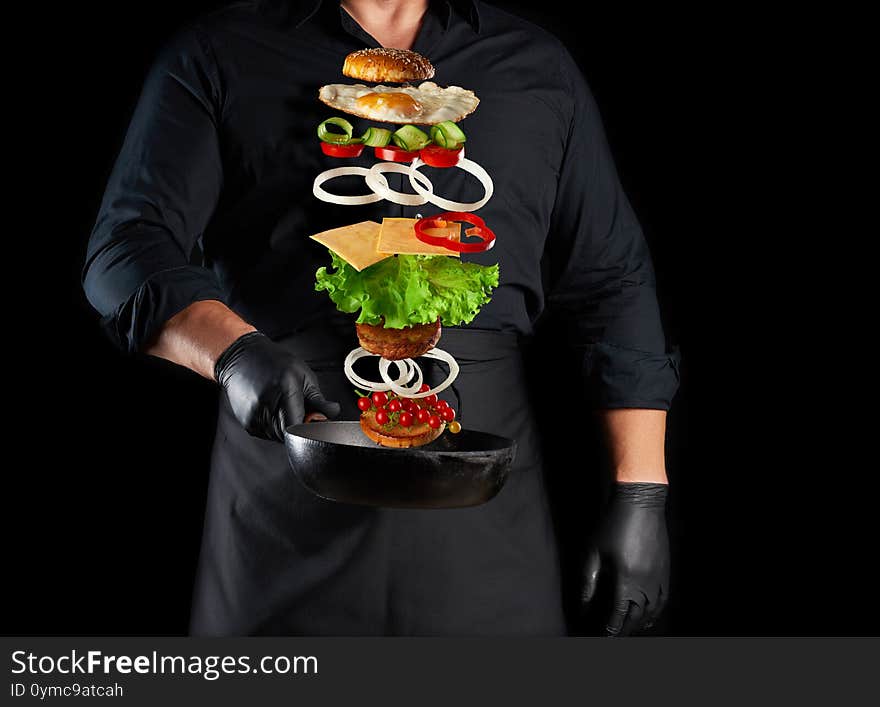 Adult Man In A Black Uniform Holding A Cast Iron Round Frying Pan With Levitating Cheeseburger Ingredients