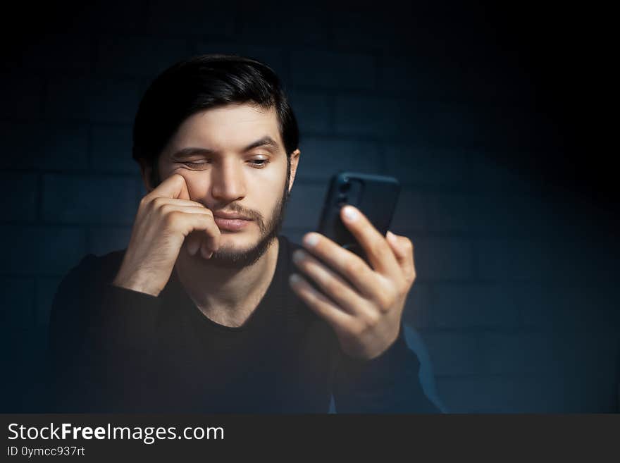 Portrait of sad young man, using smartphone, on the background of black brick wall.