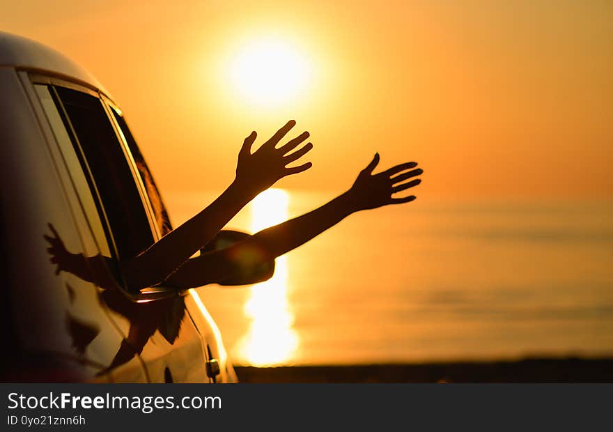 The car is on the beach at sunset. Two hands of travelers waving against the background of the sun and the sea. The concept of a family road trip at sea