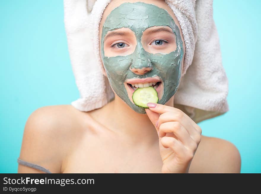 Positive smiling caucasian girl with clay cosmetic mask and organic cucumber slices, spa woman caring for her skin and health, eating organic food, isolated over blue background. Positive smiling caucasian girl with clay cosmetic mask and organic cucumber slices, spa woman caring for her skin and health, eating organic food, isolated over blue background