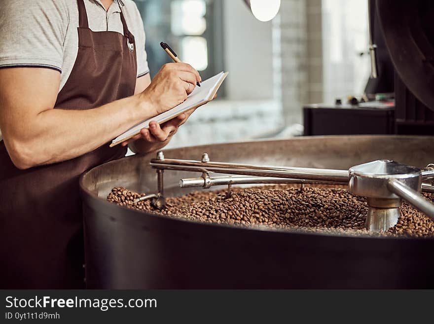 Male worker taking notes while controlling work of coffee roasting machine