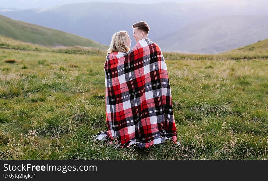 Loving Couple Sitting On A Mountain Meadow And Enjoys The View Of The Sunset