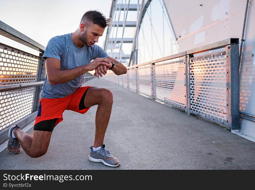 Young handsome sporty jogger taking break from exercising outdoors looking on a smart fitness watch. Sports man runner with smart watch in start position preparing to run