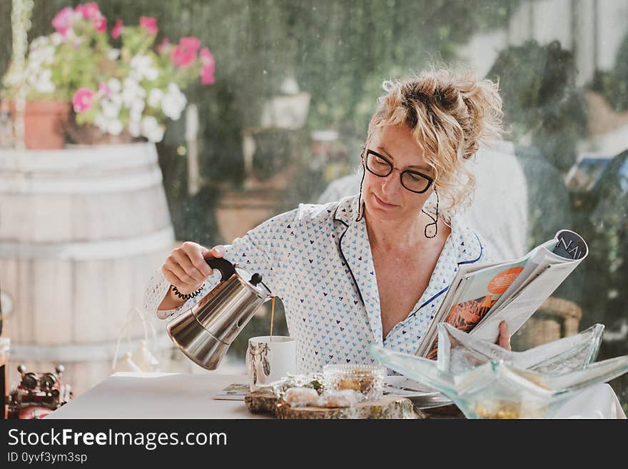 Blonde young mature woman with glasses in pyjamas at home in breakfast time, reading a magazine and preparing a cup of coffee. Blonde young mature woman with glasses in pyjamas at home in breakfast time, reading a magazine and preparing a cup of coffee