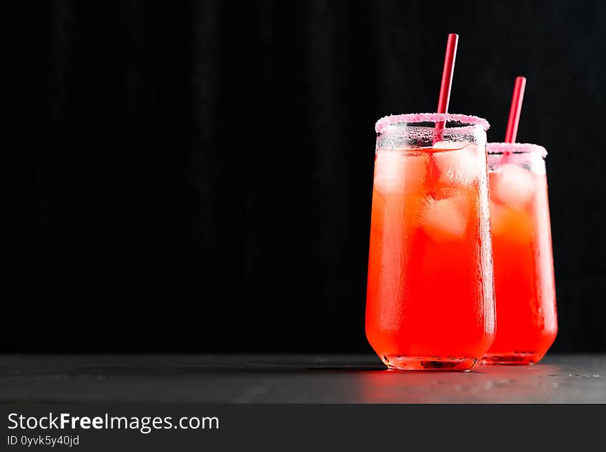 Refreshing summer mocktails with ice cubes. Two tall glasses of iced red juice with straws on dark background.