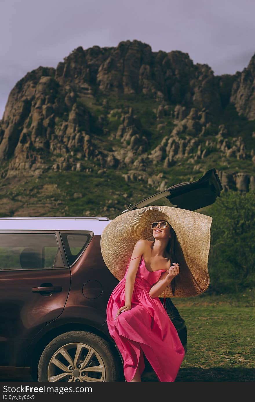 Young beautiful woman in a dress and a wide-brimmed hat near a hatchback car on a background of a mountain landscape.