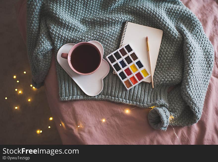 Cup of fresh coffee on plate with palette of paints, sketch book and knitted sweater over glowing Christmas lights at background in bed closeup. Winter season. Creative concept. Top view. Cup of fresh coffee on plate with palette of paints, sketch book and knitted sweater over glowing Christmas lights at background in bed closeup. Winter season. Creative concept. Top view