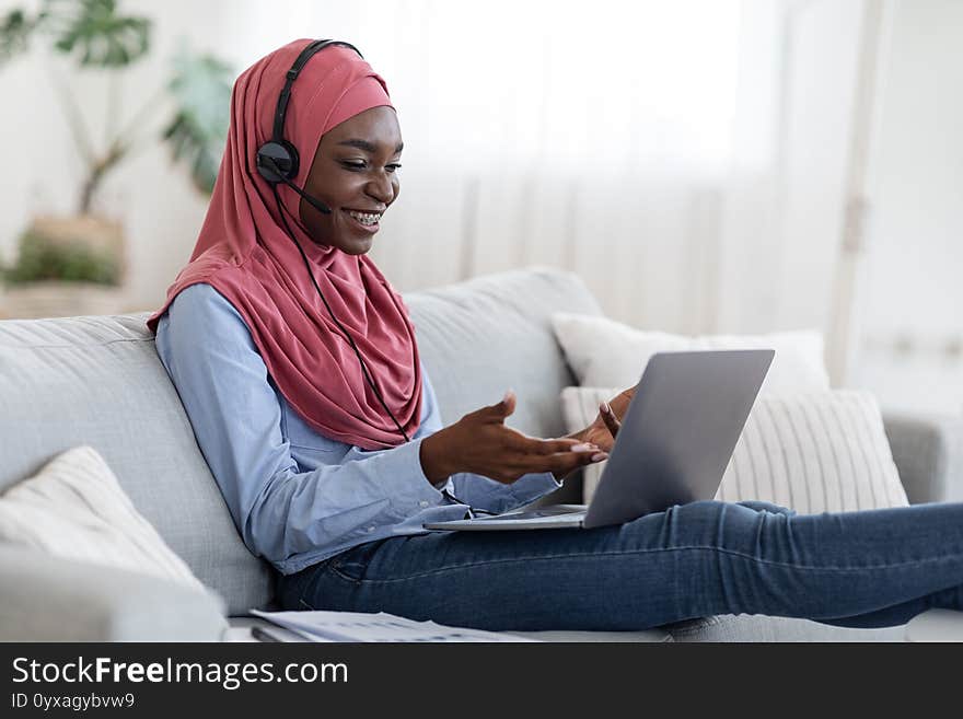 Online business meeting. Smiling black muslim lady in hijab using headset and laptop at home, having video conference, sitting with computer on comfortable couch in living room, talking on pc camera. Online business meeting. Smiling black muslim lady in hijab using headset and laptop at home, having video conference, sitting with computer on comfortable couch in living room, talking on pc camera