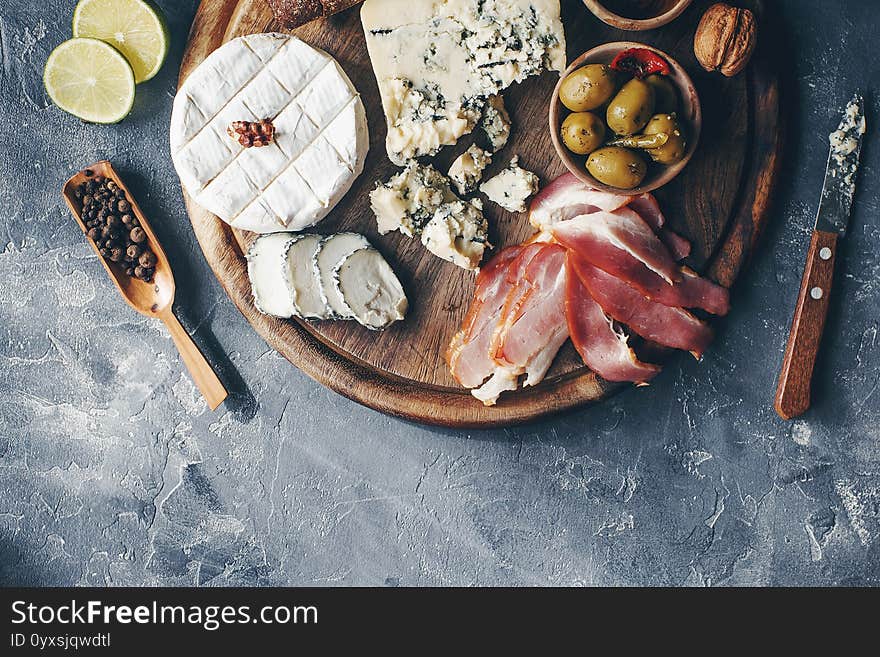 Set of cheese, hamon, honey, green olives, nuts and spices on rustic wooden board, variety of snacks on dark stone table, top view