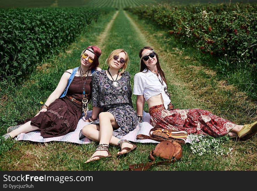 Three Hippie Women, Wearing Colorful Boho Style Clothes, Sitting, Relaxing On Green Grass Of Currant Field On Sunny Summer Day.