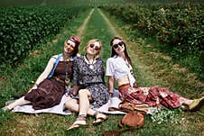 Three Hippie Women, Wearing Colorful Boho Style Clothes, Sitting, Relaxing On Green Grass Of Currant Field On Sunny Summer Day. Stock Images