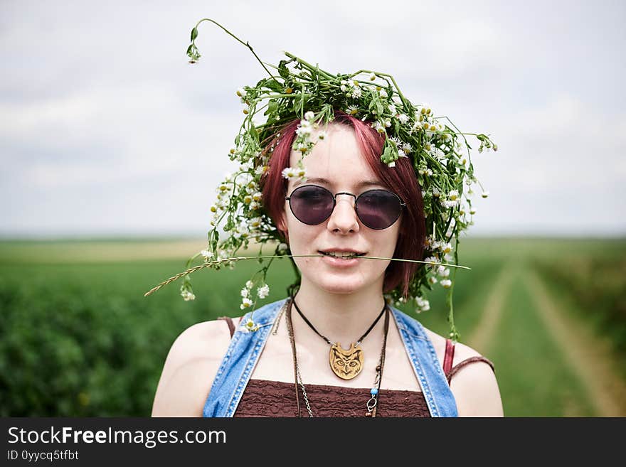 Young hippie woman with short red hair, wearing boho style clothes and flower wreath, standing on green currant field, posing for