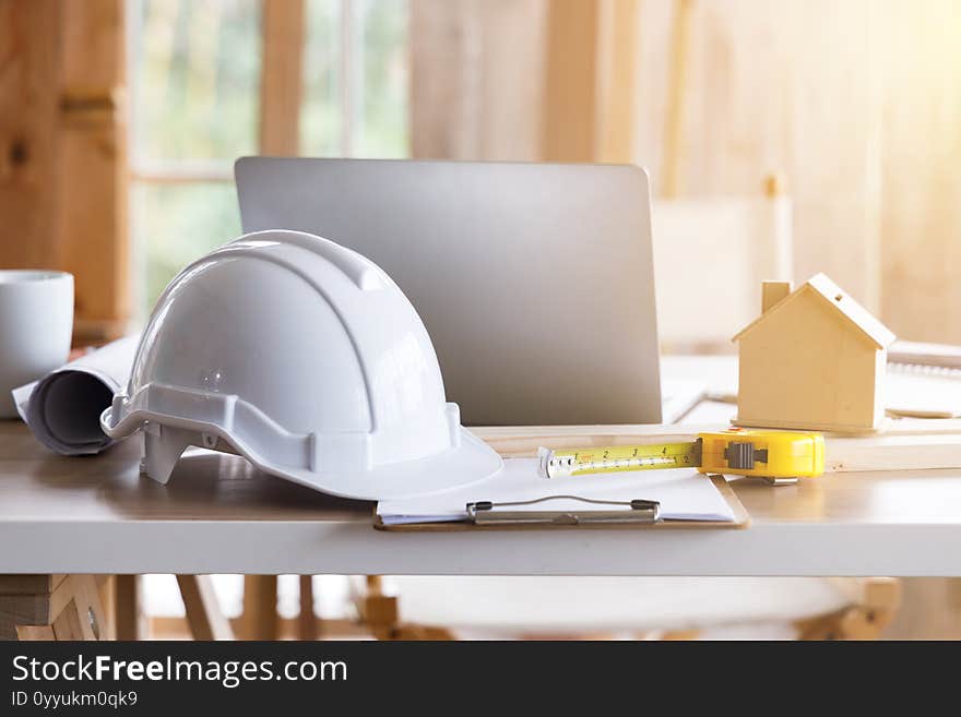 White safety helmet on messy engineer`s table with tape measure, tiny wooden house model and laptop computer. White safety helmet on messy engineer`s table with tape measure, tiny wooden house model and laptop computer.